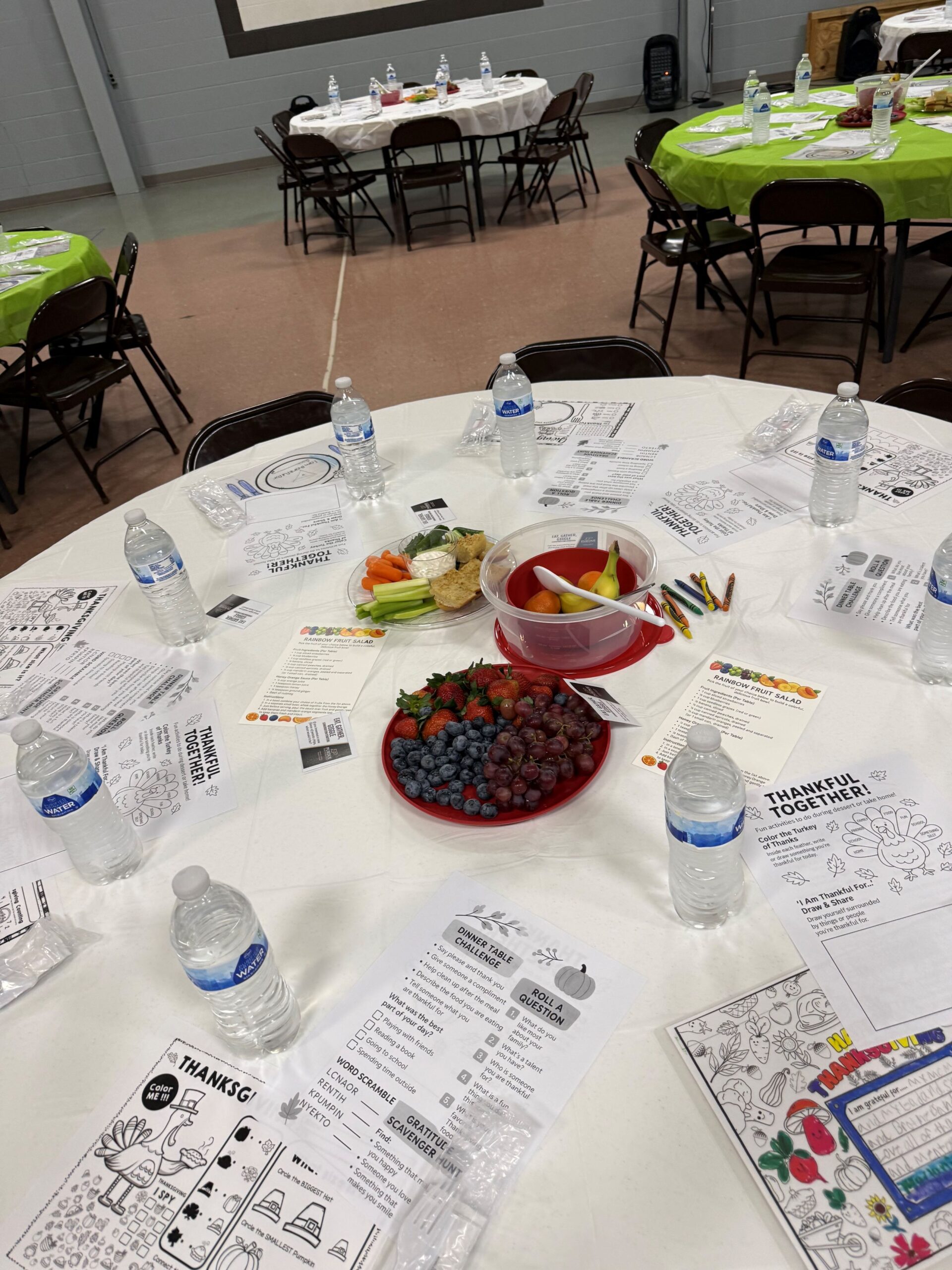 table set with water bottles, worksheets, in the center is bowl of fruit, plate and veggies. In background is three other tables set the same in a gym setting.